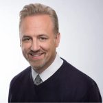 A smiling man with light hair and a goatee, wearing a dark sweater over a collared shirt, against a light background.